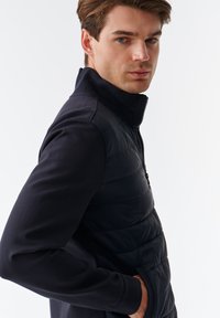 Marineblå quiltet pufferjakke med en høj krave, halv-zip lukning og lange, tætsiddende ærmer. Glat tekstur og mat finish.