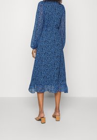 Modrá květinová midi šaty s dlouhými rukávy, průhlednou látkou, volánkovým lemem a vymezeným pasem. Nosené s béžovými muly na blokovém podpatku.