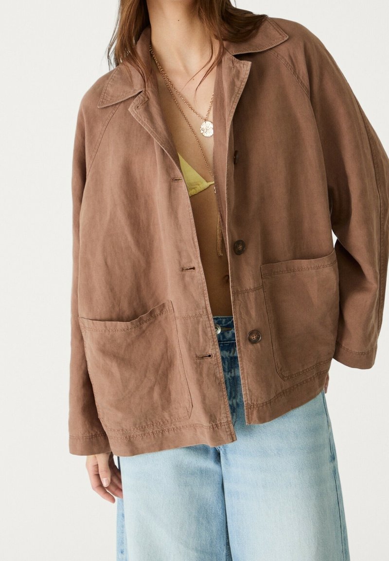 Personne portant une veste marron boutonnée oversize, un haut jaune, des colliers en or superposés et un jean bleu clair sur un fond uni.