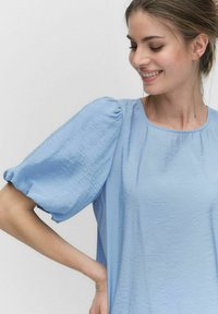 Lys blå bluse med pufset korte ærmer, lavet af struktureret stof. Rundt udskæringer og løs pasform, med minimale design elementer.