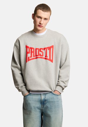 Młody mężczyzna z krótkimi włosami ubrany w jasnoszarą bluzę z wyraźnym czerwonym napisem "PROSTO" oraz jasnoniebieskie jeansy, ręce w kieszeniach.