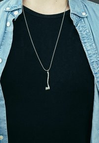 Collana con pendente d'argento a forma di ascia, pendente da una catena. Indossata su una maglietta nera, incorniciata da una giacca di jeans blu chiaro.