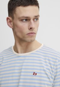 Juostelėta trumpomis rankovėmis t-shirt šviesiai mėlynos ir kreminės spalvų, su apvaliu kaklo iškirpimu ir mažu raudonu logotipu krūtinėje. Lygus medvilnės paviršius.