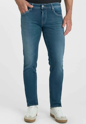 Mann, der eng anliegende blaue Jeans und weiße Sneakers trägt, steht mit einer Hand in der Tasche vor einem einfarbigen Hintergrund.