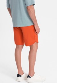 Shorts de sport orange à coupe décontractée, avec une texture lisse et des coutures contrastantes, associés à un T-shirt bleu clair et des baskets blanches.