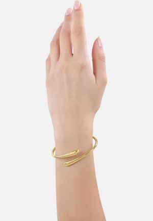 Bracciale in metallo color oro, liscio con un design minimalista, caratterizzato da una forma aperta e curve morbide. Indossato su un polso sottile.
