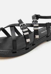 Sandali neri con cinturini sintetici testurizzati che presentano accenti quadrati in argento, una suola piatta e un design open-toe.