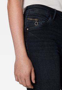 Tummansinisen denim-kankaan slim fit -farkut, joissa on vetoketjullinen_tasku kultaisilla yksityiskohdilla ja metallinen korostus vetoketjussa.