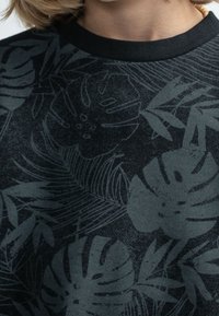 Černá mikina s tmavě zeleným potiskem tropických listů, žebrovaným výstřihem a texturovanou tkaninou, která kombinuje květinové a listové vzory.