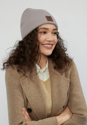 El gorro de punto gris presenta una textura acanalada y una etiqueta de cuero marrón. El modelo lleva un abrigo beige sobre un suéter claro con una camisa de cuello.