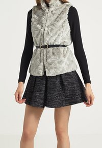 Faux fur-väst i ljusgrått med hög krage och bälte i midjan, parat med en svart texturerad kjol med en utställd design.