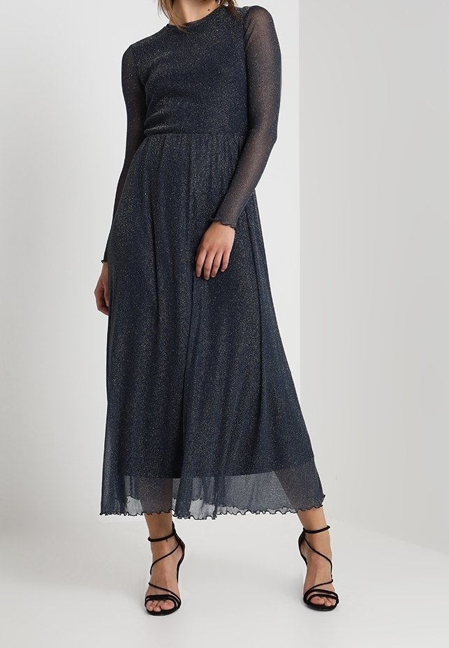 Robe à manches longues, bleu foncé, en matière translucide et scintillante. Présente un corsage ajusté et une jupe fluide avec un ourlet en dents de scie, assortie à des chaussures à talons noires.