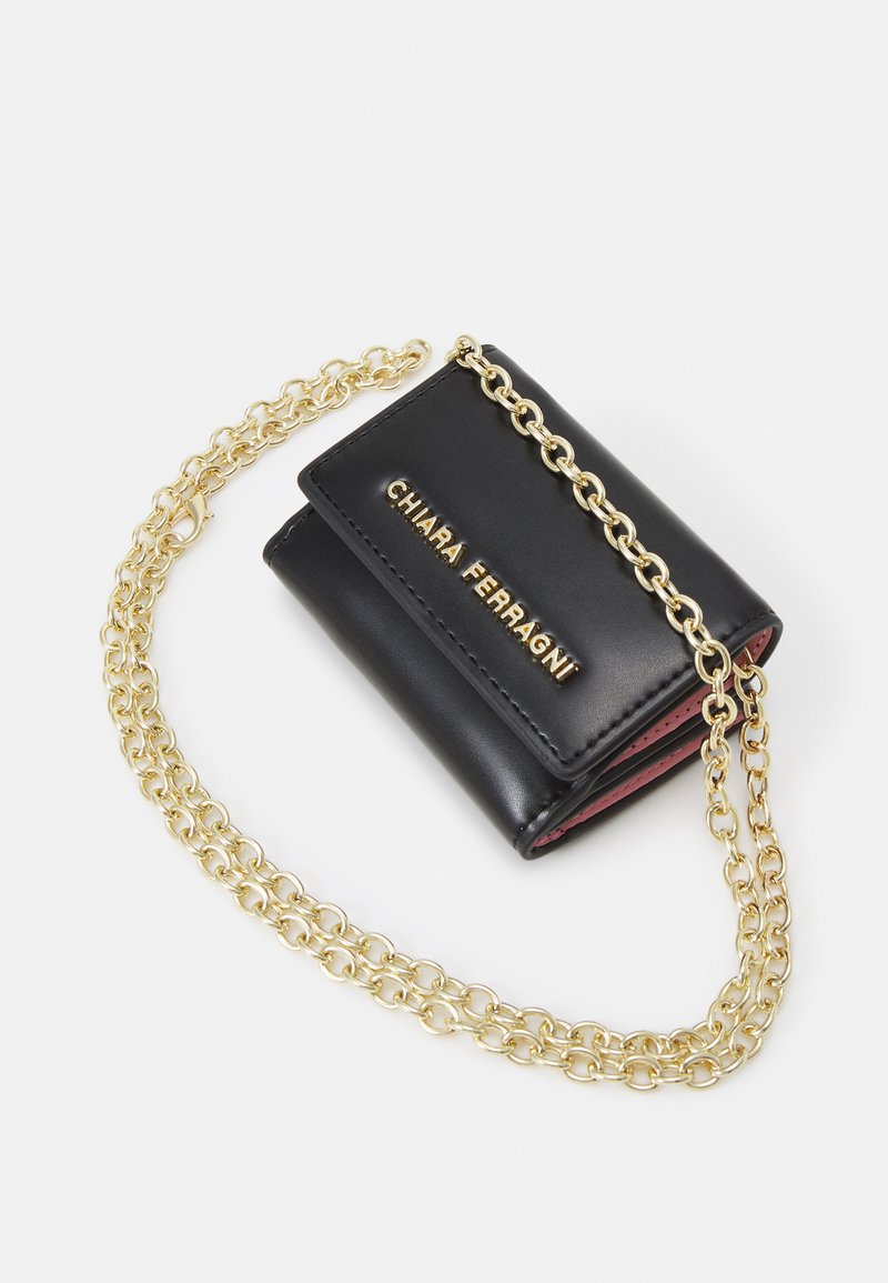 Portefeuille en cuir noir avec une chaîne dorée et le logo "Chiara Ferragni" en or à l'avant, bords intérieurs roses visibles, sur fond blanc.