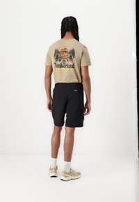 Beige tričko s barevným grafickým motivem Timberland; spárováno s černými šortkami a béžovými atletickými botami s bílými ponožkami.