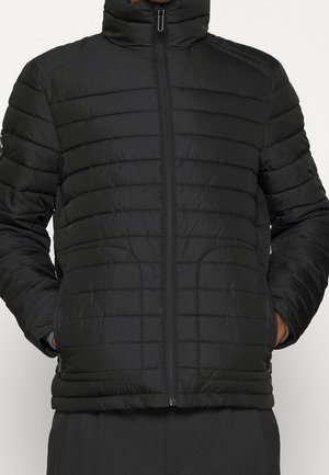 Sort quiltet jakke med vandrette syninger, frontlynlås og hænder i sidelommer, båret af en person mod en ensfarvet baggrund.