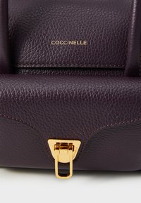 Sac à main en cuir texturé violet avec un fermoir doré et un logo de marque embossé, présentant des détails de couture visibles.