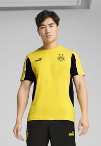 Kollane lühikeste varukate spordisärk mustade külgedega paneelidega, esitades Puma logo ja Borussia Dortmundi graafika ees. Sile kangatekstuur.