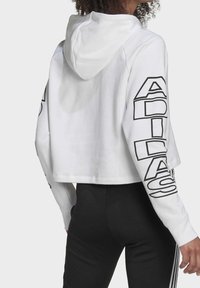 White cropped hoodie with long sleeves featuring black "ADIDAS" text along one arm. Smooth fabric, with a hood and relaxed fit.