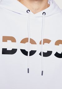 Sudadera blanca con la palabra "BOSS" en letras gruesas. Las letras están en tonalidades de marrón, negro y blanco. Incluye cordones con puntas metálicas.