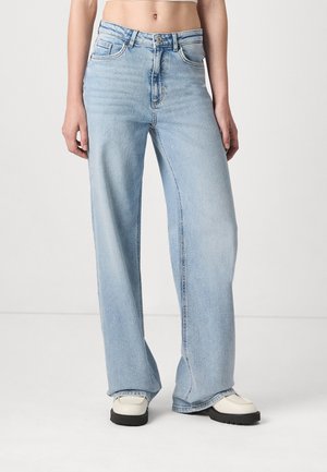 Persoon die lichtblauwe, hooggetailleerde jeans met wijde pijpen en witte platformschoenen draagt, staande tegen een effen witte achtergrond.
