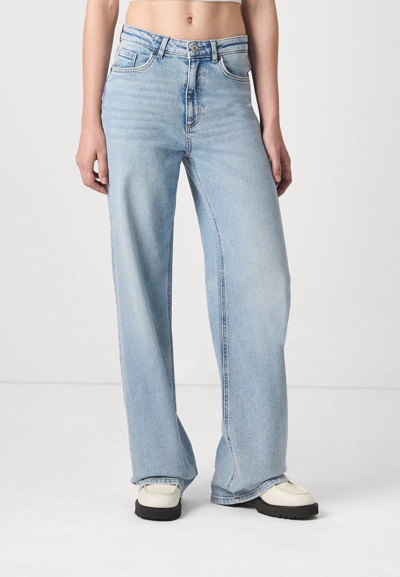 Personne portant un jean bleu clair taille haute à jambes larges et des chaussures compensées blanches, debout devant un fond blanc uni.