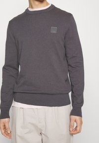 Mężczyzna w ciemnoszarym swetrze z okrągłym dekoltem, pod który ma jasnoróżową koszulę, oraz beżowych spodniach, stojący na jednolitym tle.