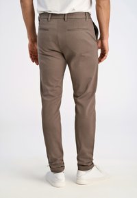 Pantaloni marroni su misura con una texture liscia, taglio slim e tasche posteriori discrete. Indossati con scarpe da ginnastica bianche.