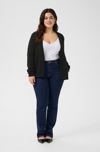 Un cardigan noir sur un haut blanc à col en V, associé à un jean bleu foncé à taille mi-haute. Le modèle se tient debout avec des escarpins noirs à bout pointu sur un fond neutre.