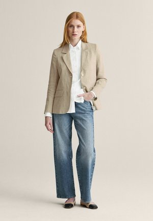 Kvinde står iført beige blazer, hvid skjorte, blå jeans og beige flade sko med sorte tåhætte på en ensfarvet lys baggrund.