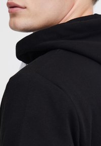 Felpa nera con cappuccio e texture liscia, caratterizzata da una vestibilità comoda e cuciture visibili lungo la linea del collo e delle spalle.