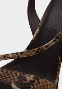 Κλείσιμο εικόνας ενός καφέ δερμάτινου σανδαλιού με λεπτά, φιδίσια σχέδια λουριά που διασταυρώνονται πάνω σε μια σκούρα καφέ εσωτερική σόλα.