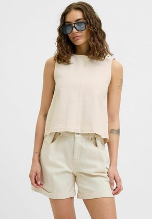 Top sin mangas de color beige con un diseño cuadrado, textura suave y cuello alto, combinado con pantalones cortos enrollados de un beige claro. Las gafas de sol acentúan el look.