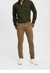 Groene button-down shirt met twee borstzakken, gecombineerd met tan slim-fit broeken. Witte sneakers maken de outfit af.