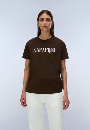 Femme aux cheveux bruns et raides portant un t-shirt marron Napapijri et un pantalon blanc, debout devant un fond clair uni.