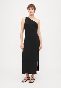 Vestido maxi negro de un solo hombro con textura acanalada, detalles de abertura lateral y diseño ajustado, combinado con sandalias minimalistas negras.