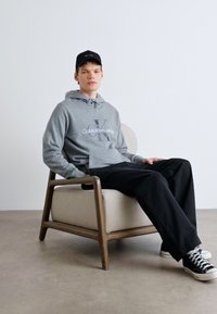 Felpa grigia con logo "Calvin Klein Jeans", pantaloni neri dal taglio rilassato e sneakers nere. Seduto su una sedia in legno minimalista.