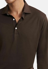 Vīrietis ģērbts tum�ši brūnā garo piedurkņu polo krekla ar divām baltām pogām, kas ir aizpogāta, un ar nolocītu apkakli, redzams no kakla līdz viduklim.