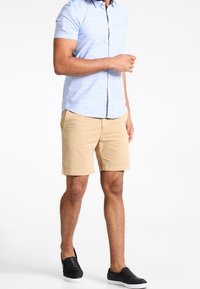 Chemise à manches courtes à carreaux bleu clair avec une poche poitrine, associée à un short beige et des chaussures noires sans lacets. Tissu lisse, coupe décontractée.