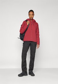 Veste rouge imperméable avec capuche, pantalon cargo noir et chaussures de randonnée noires. La personne tient un sac �à dos noir. Design simple, tissu léger.