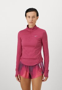 Nike lange mouw top in roze stof met een half-zip ontwerp, voorzien van reflecterende accenten. Gecombineerd met multikleurige, verloop korte broek.