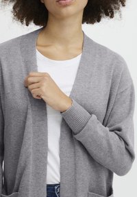 Šedý pletený cardigan s žebrovanými manžetami a otevřeným předním dílem, v kombinaci s bílým tričkem. Má jemnou texturu a ležérní design.