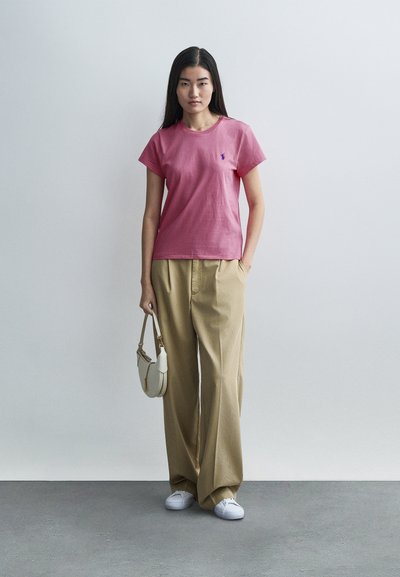 Rózsaszín rövid ujjú póló kis logóval, bő bézs nadrággal és fehér tornacipővel párosítva, kezében egy kis krémszínű kézitáska.