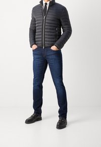 Svart vadderad jacka med horisontell sömnad, blå jeans med lätt blekning och svarta loafers med gulddetaljer.