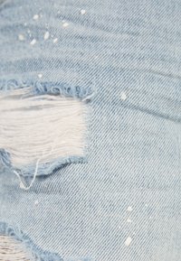 Ljust blå denimtyg med fransiga rivningar och vita färgstänk, som visar en sliten, urblekt textur och slitna trådar.