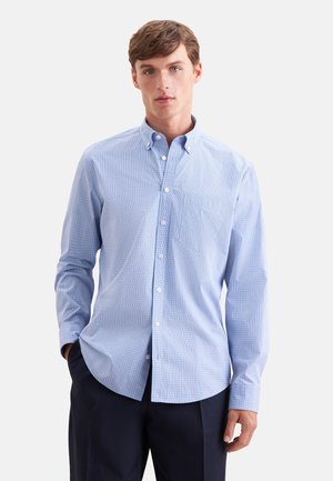 Junger Mann trägt ein hellblau kariertes Hemd mit Knöpfen und langen Ärmeln und dunkle Hose, steht mit einer Hand in der Tasche.
