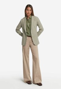 Ljusgrå texturerad kavaj med slag, kombinerad med en grön skjorta med knappar och beige utsvängda byxor. Bruna flätade sandaler fulländar looken.