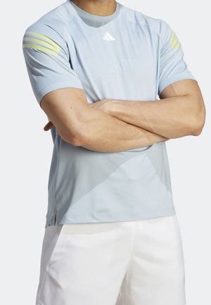 Hombre con los brazos cruzados que lleva una camiseta deportiva azul claro de manga corta con rayas amarillas en las mangas y pantalones cortos blancos.