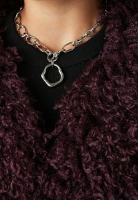 Collana chunky in argento con pendente geometrico irregolare indossata sopra una maglietta nera e un capo in tessuto riccio scuro di colore viola.