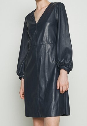 Kobieta w czarnej sukience z ekoskóry z długimi bufkami na rękawach i dekoltem w serek, pokazana od ramion do połowy ud na jednolitym tle.