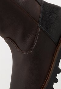 Ruskea nahkasaapas, jossa on teksturoitu pinta, kontrastiväriset mustat yksityiskohdat, harmaa ompelu ja kova pohja, jossa on havaittavat urat.